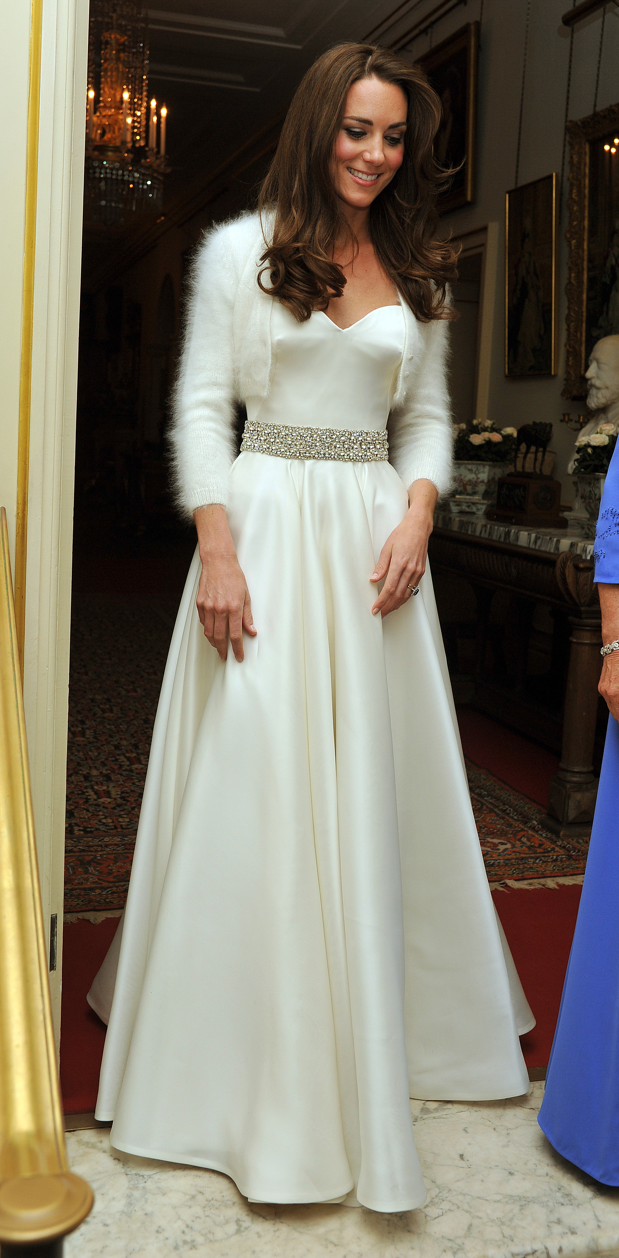 Kate Middleton wearing a long silky white a-line dress with a sparkly belt and a fluffy white cardigan 