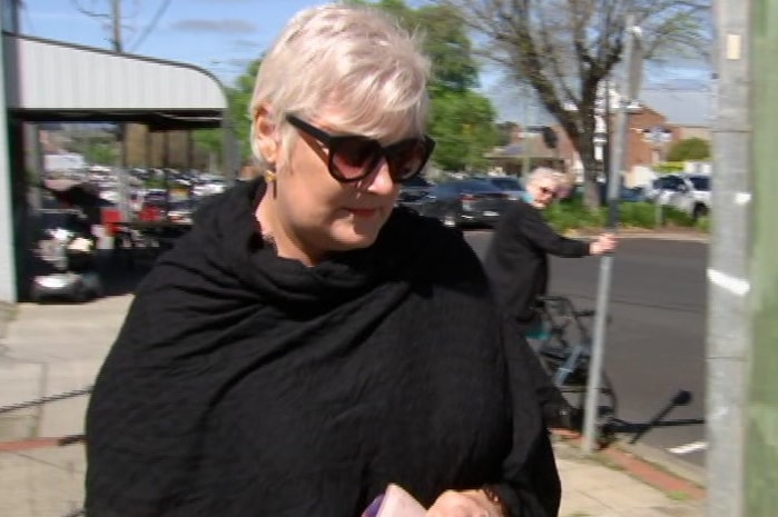A woman with short, grey hair, wearing a black shawl-like top and sun glasses walks along the street.