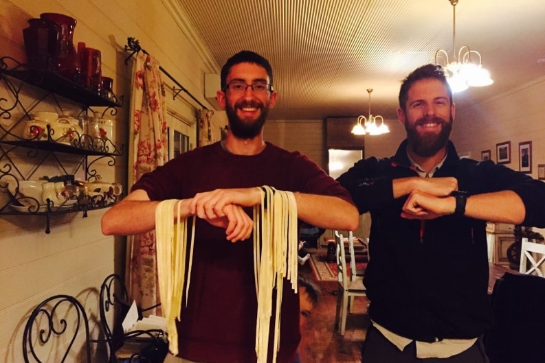 Two men with beards stand inside a kitchen smiling, strips of dry spaghetti hanging from their arms.