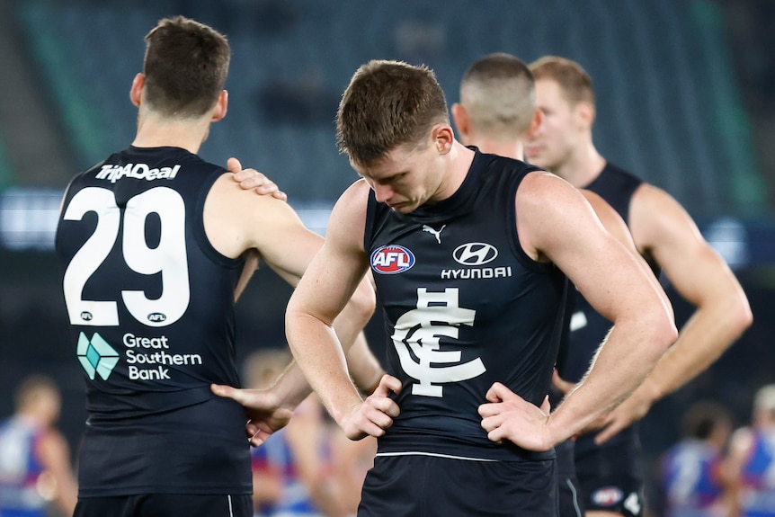 A Carlton AFL player looks dejected with his teammates standing behind him after a loss to the Western Bulldogs.