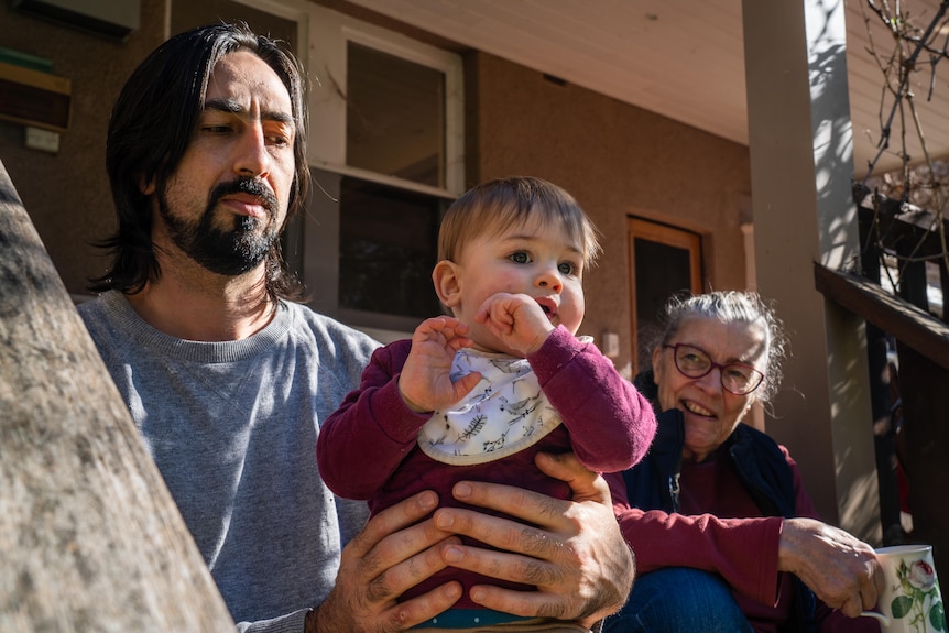 A man, his mother in law and young child sit on the back step of their house in the sunshine.