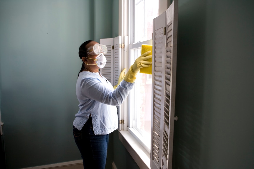 A woman wears a mask and goggles wiping a window with a sponge.