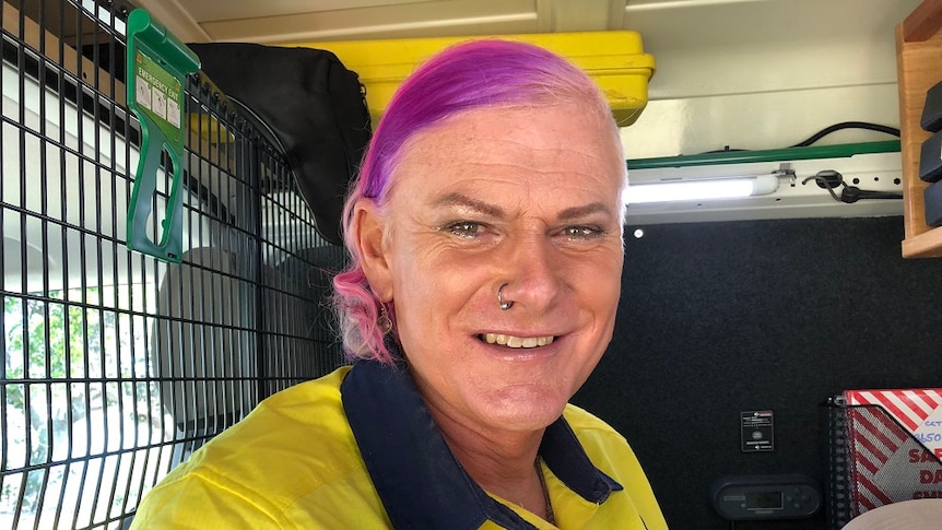 A man dressed as a woman with purple hair and an earring in their nose sitting inside a cabin of a work truck