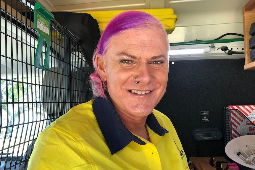 A man dressed as a woman with purple hair and an earring in their nose sitting inside a cabin of a work truck