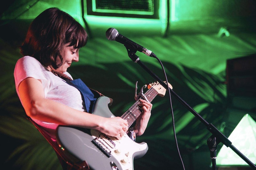 Stella Donnelly smiles while standing at a microphone and playing guitar
