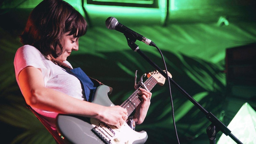 Stella Donnelly smiles while standing at a microphone and playing guitar