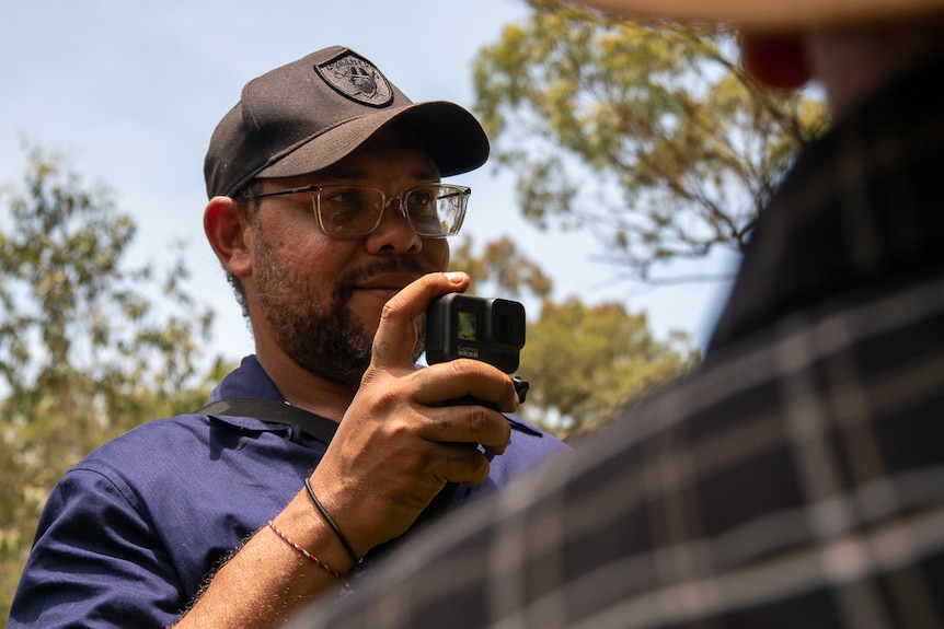 Young Aboriginal man filming Elder with GoPro while searching for bush food