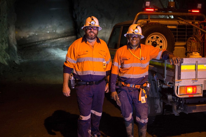 two men in hi-vis clothing and hard hats standing next to a ute underground.