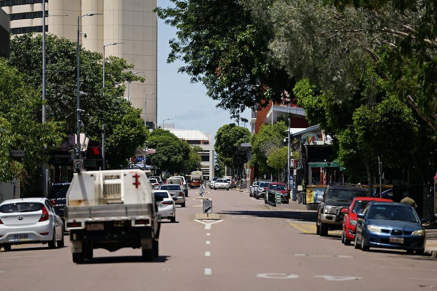 A double-lane street lined with trees and a tall building on the left. A ute is driving away