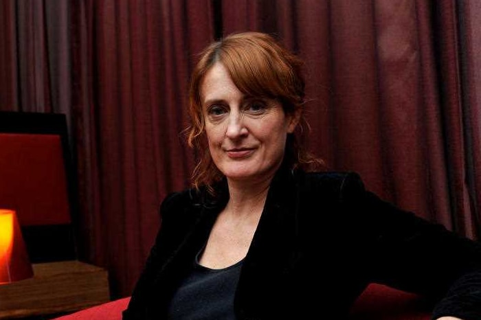 A woman with red hair is sitting down with a red velvet backdrop