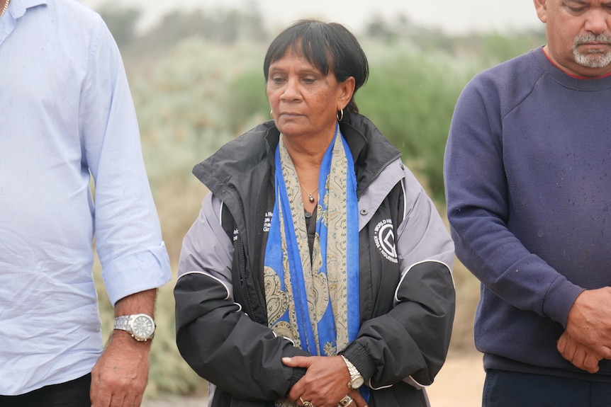 Woman with short black hair, eyes downcast, wearing a large black and purple sports jacket, standing between two much taller men