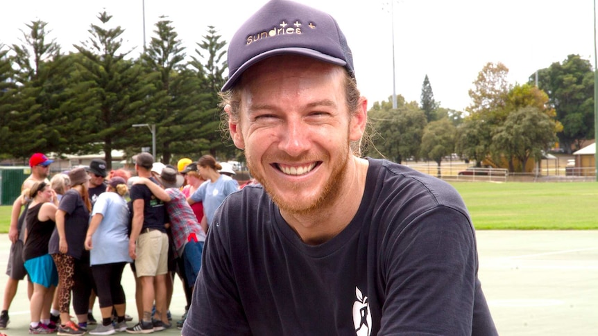 A young smiling man in a cap with a group of people in the background in a team huddle.