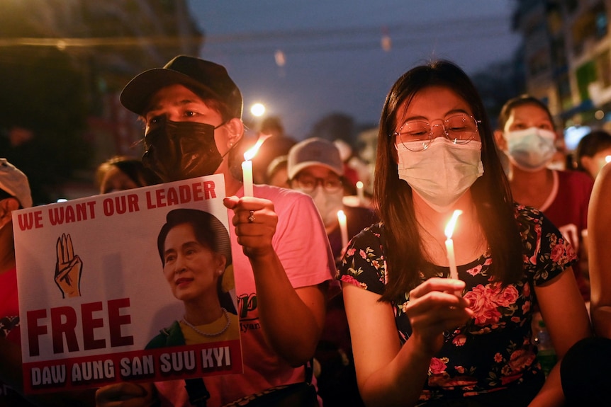 A woman in a mask holding a candle next to another protester with a sign depicting Aung San Suu Kyi
