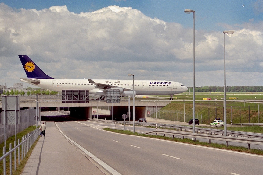 You view a near-empty dual carriageway which leads to an airport runway overpass with a large Lufthansa plane taxiing.
