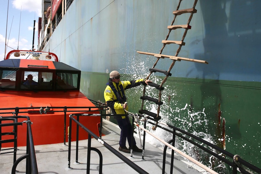 A man stands on a small boat next to a tanker ship with a rope ladder on the side.