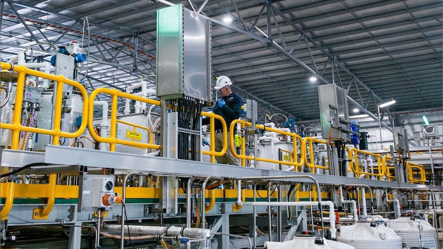 A man with a helmet on is standing inside a processing plant.
