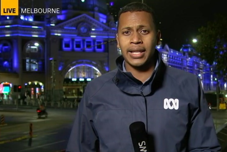 Clure holding microphone standing in front of Flinders Street Station in dark.