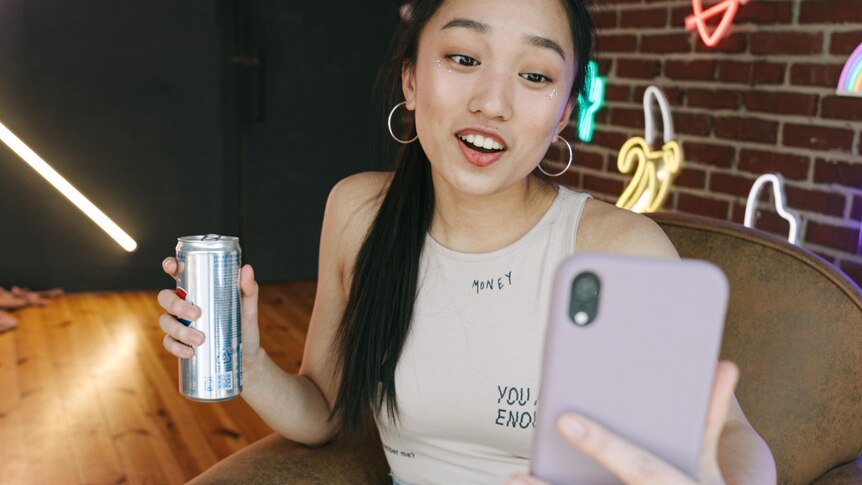 Photo of a young woman holding a can of drink taking a selfie against a brick wall adorned with neon logos.