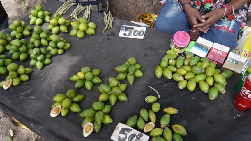 piles of green betel nut sit on a black table cloth covered street table for sale alongside cigerettes and fizzy drink cans. 
