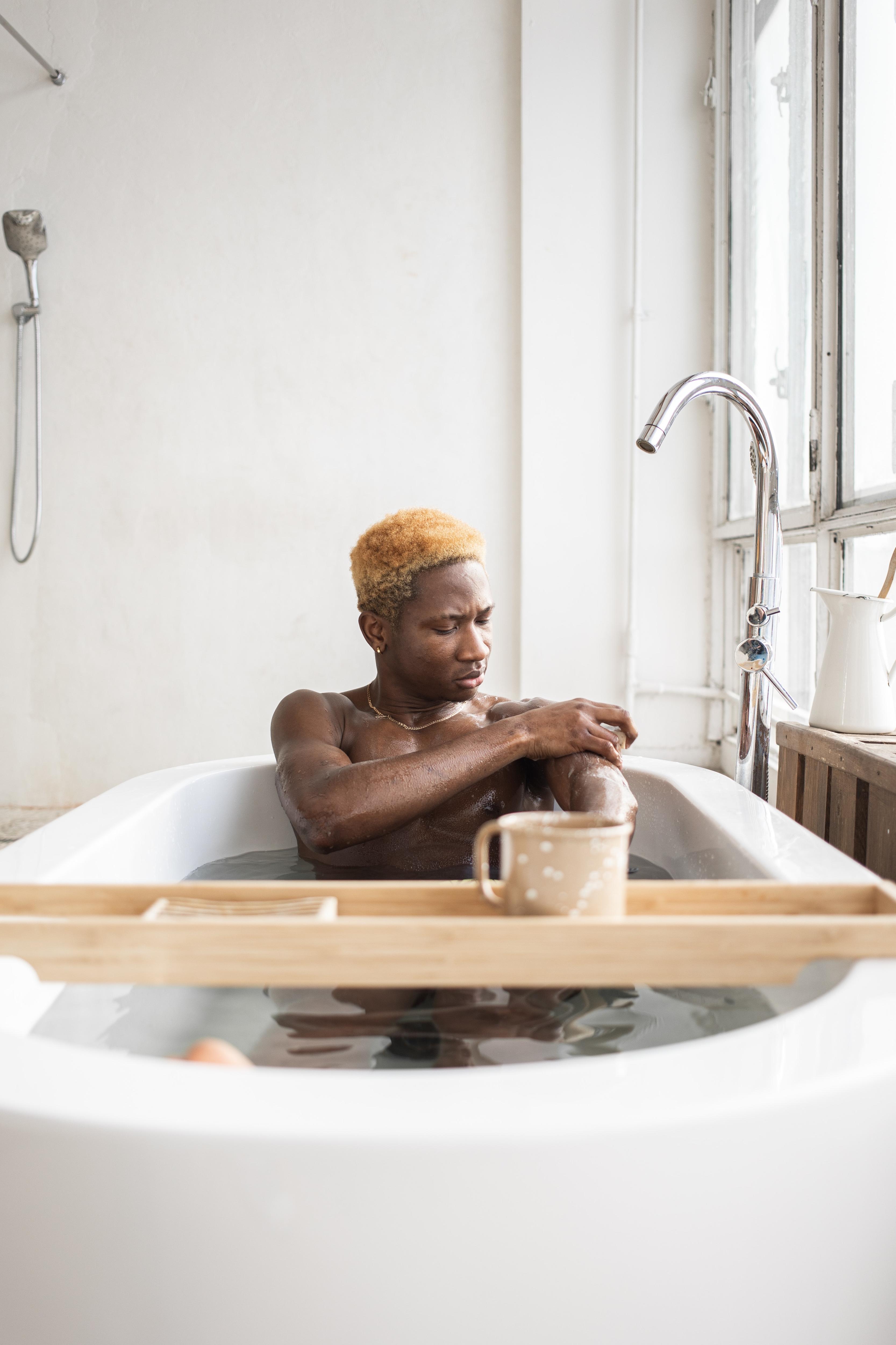 A man with dark skin is sitting in a white bathtub, washing his left arm.