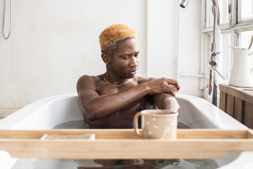 A man with dark skin is sitting in a white bathtub, washing his left arm.