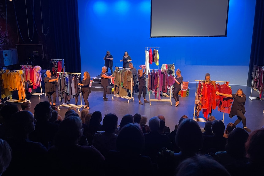 A group of women sorting through clothes racks on stage perform to a full audience  