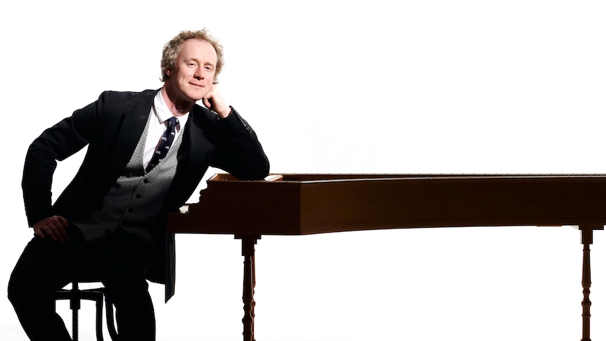 Harpsichordist Donald Nicolson sitting at a keyboard leaning against it, dressed in a black suit with a white shirt and a tie.