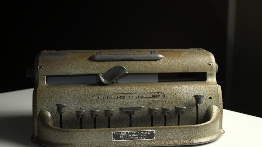 A Perkins Brailler sits on a table. It is a grey cast iron braille typewriter with nine levers at the front.