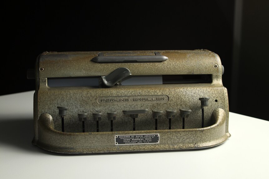 A Perkins Brailler sits on a table. It is a grey cast iron braille typewriter with nine levers at the front.