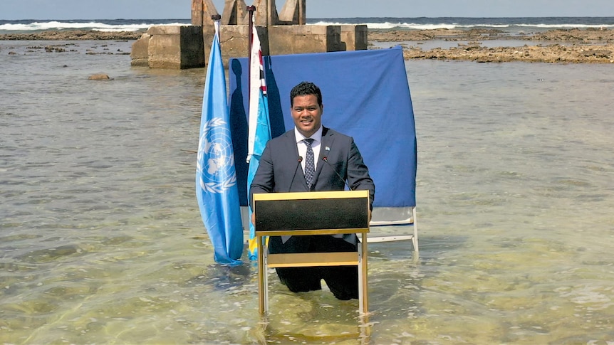 Man in suit and tie stands before a blue sheet and two flags, up to his knees in water, with ocean surrounding him.