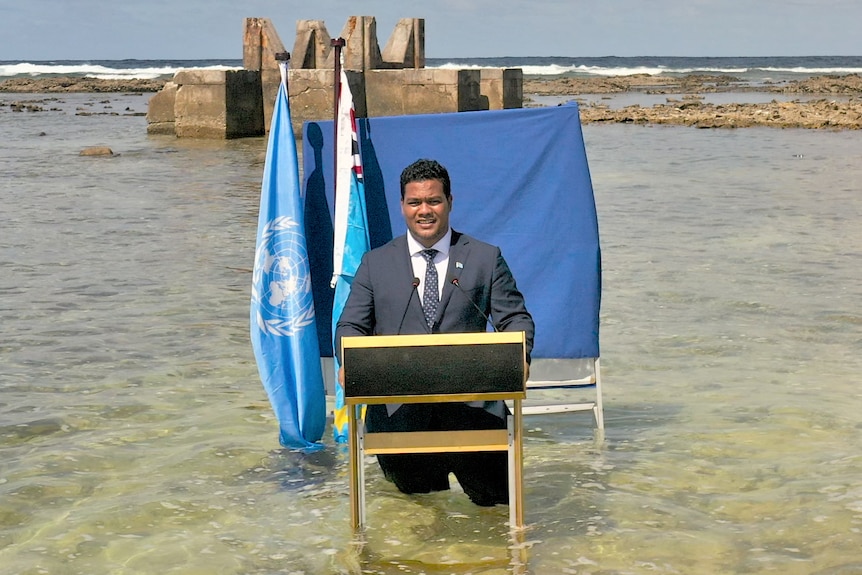 Man in suit and tie stands before a blue sheet and two flags, up to his knees in water, with ocean surrounding him.