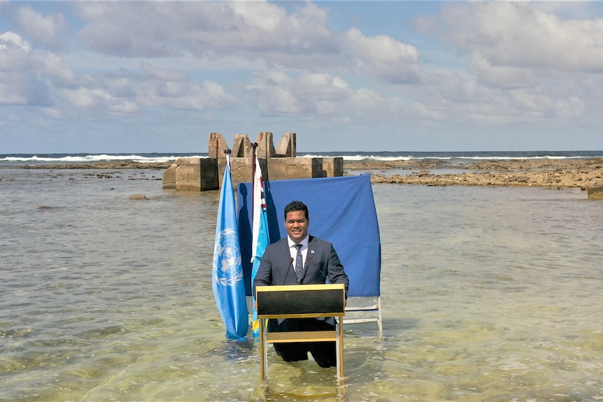 Man in suit and tie stands before a blue sheet and two flags, up to his knees in water, with ocean surrounding him.