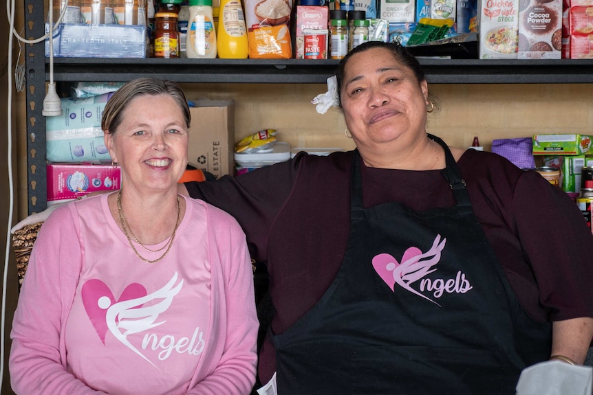 Two women sit, smiling, in front of a shelf of food in packets.