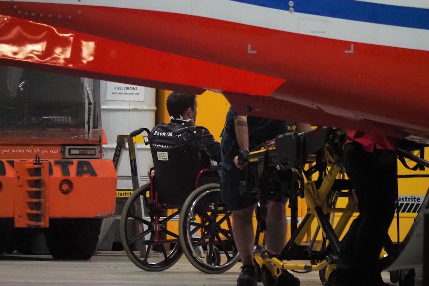 A man sits in a wheelchair while paramedics handle a stretcher nearby.