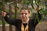 A woman dances at an Aboriginal smoking ceremony. She has traditional makeup and jewellery and holds branches