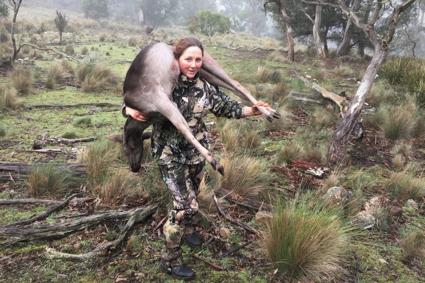 A woman looking pleased as she carries a dead deer over her shoulders through bushland.