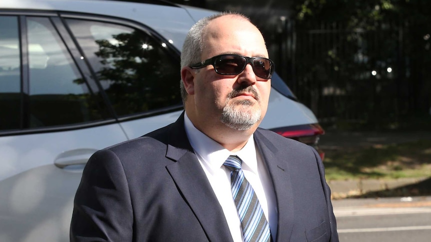 A middle-aged man wearing a suit and sunglasses walks towards the Coroners Court of Victoria.