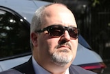 A middle-aged man wearing a suit and sunglasses walks towards the Coroners Court of Victoria.