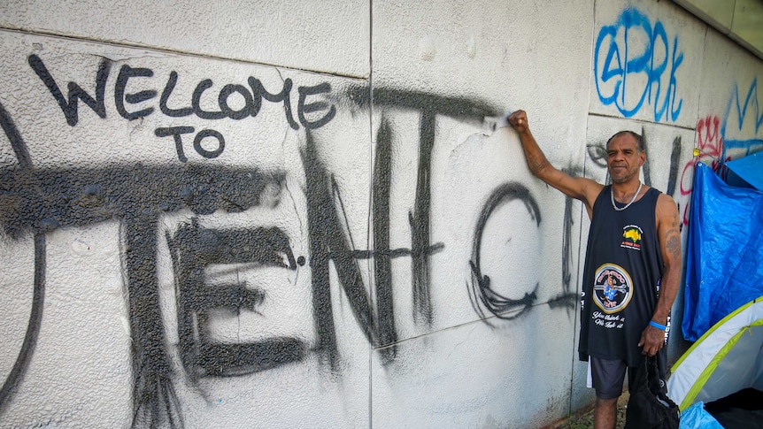 Neville Riley stands next to graffiti that reads 'Welcome to Tent City'