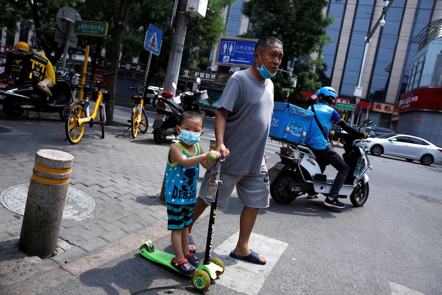 A little boy on a face mask on a scooter next to an older man in a face mask