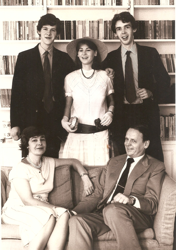Two young men in suits stand on either side of a young woman in a white dress, behind an older man and woman seated on a sofa.