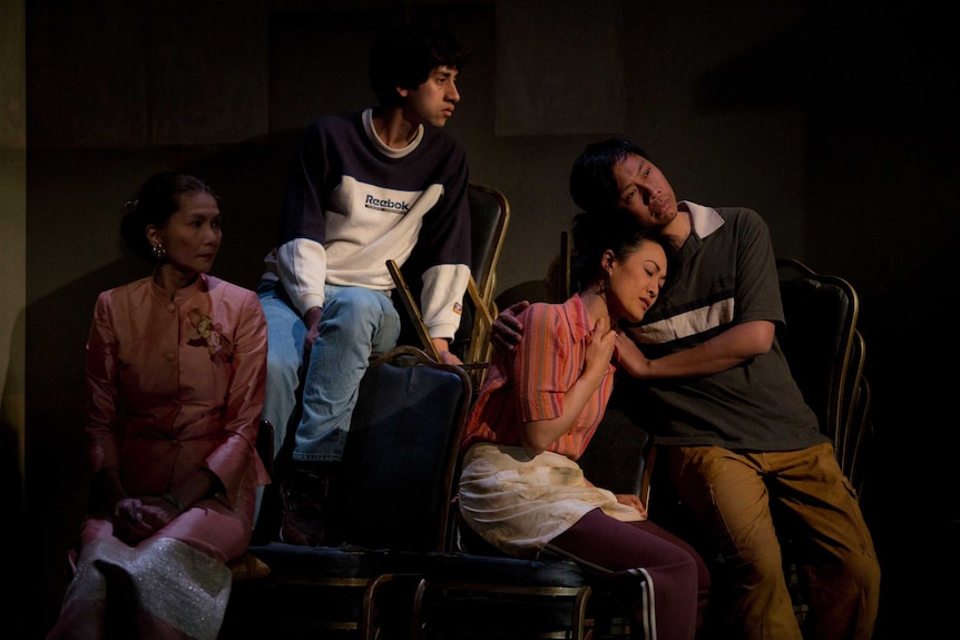 Four people seated on chairs: a serious looking woman with her son, and a couple embracing.