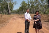 A man and a woman stand in a dirt road surround by scrub
