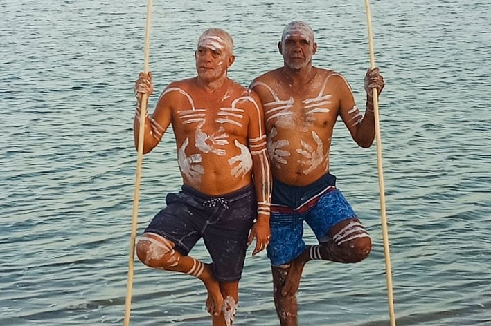 Two men in traditional ochre holding spears on beach