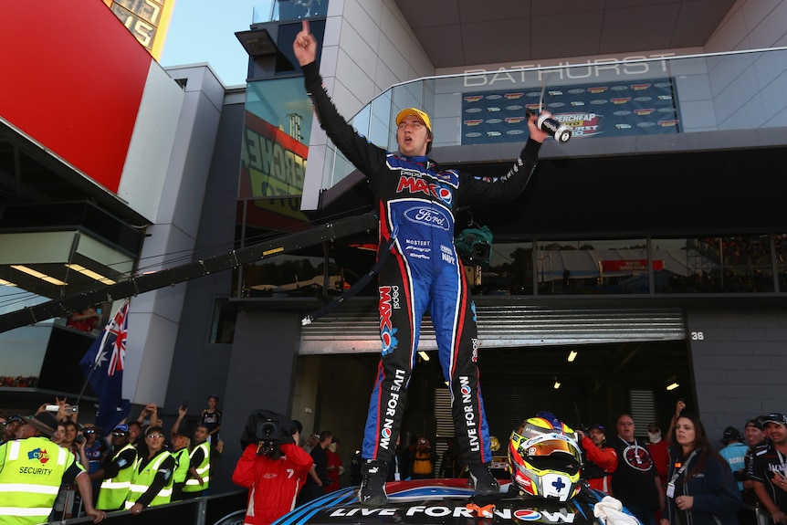 A Ford racing driver strikes a pose standing triumphant on the roof of his car after a big win.  
