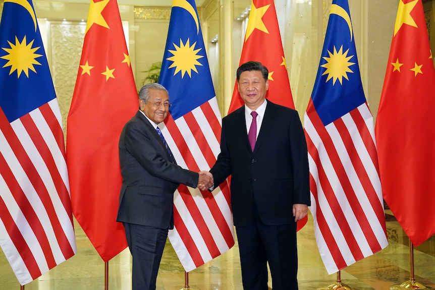 Xi Jinping shakes hands with Malaysian Prime Minister Mahathir Mohamad while standing in front of Malaysian and Chinese flags.