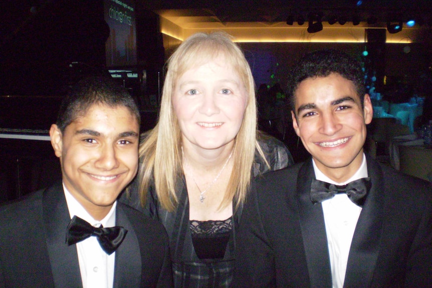 Two teenage boys wearing suits and sitting next to their mother, who is smiling in the middle.