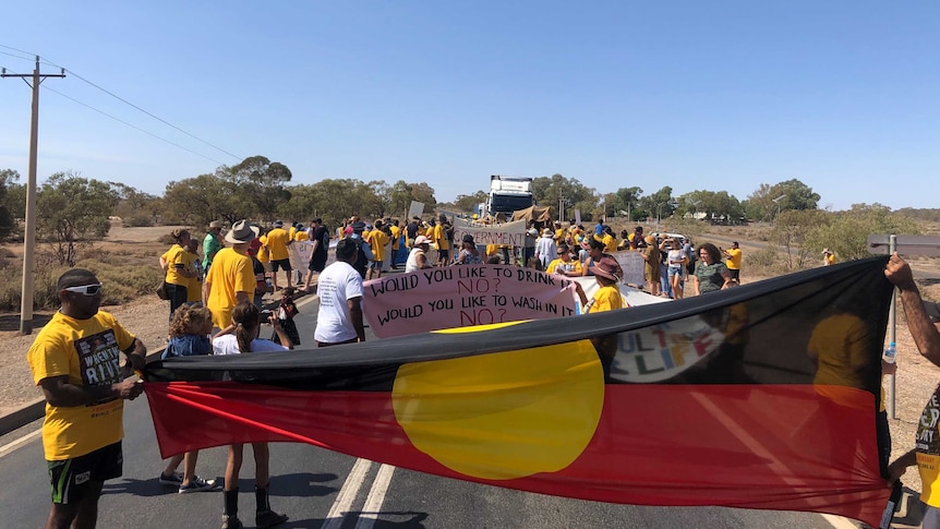 A large Aboriginal flag is stretched out in front of a crowd holding signs, blocking the way of a truck.