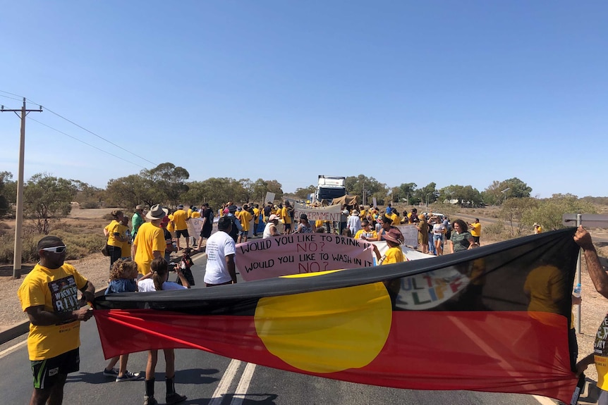 A large Aboriginal flag is stretched out in front of a crowd holding signs, blocking the way of a truck.