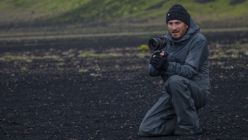 A white man in his early 50s - the director Darren Aronofsky - crouching with a camera in a dark field